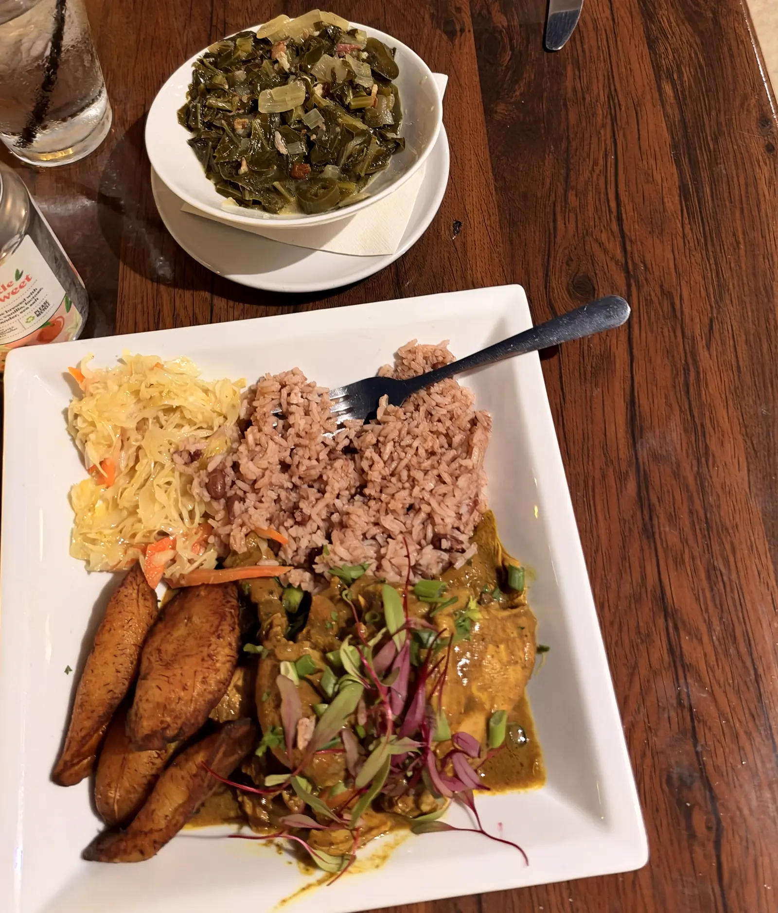 Curry chicken with rice and peas, fried plantains and slaw, beside a bowl of collard greens